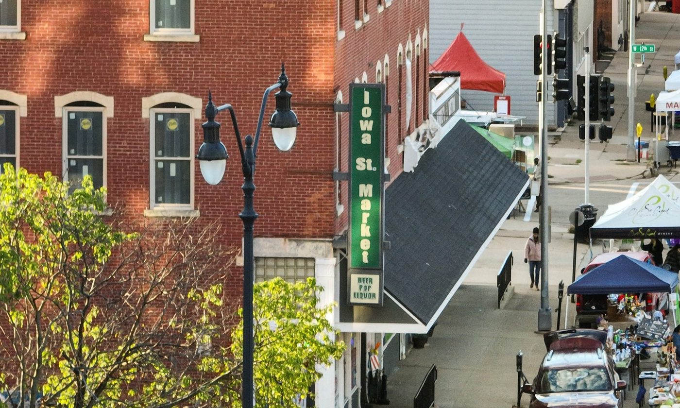 Street-level view of the Iowa St. Market sign on Iowa Street, with the Dubuque Farmers' Market set up along the sidewalk